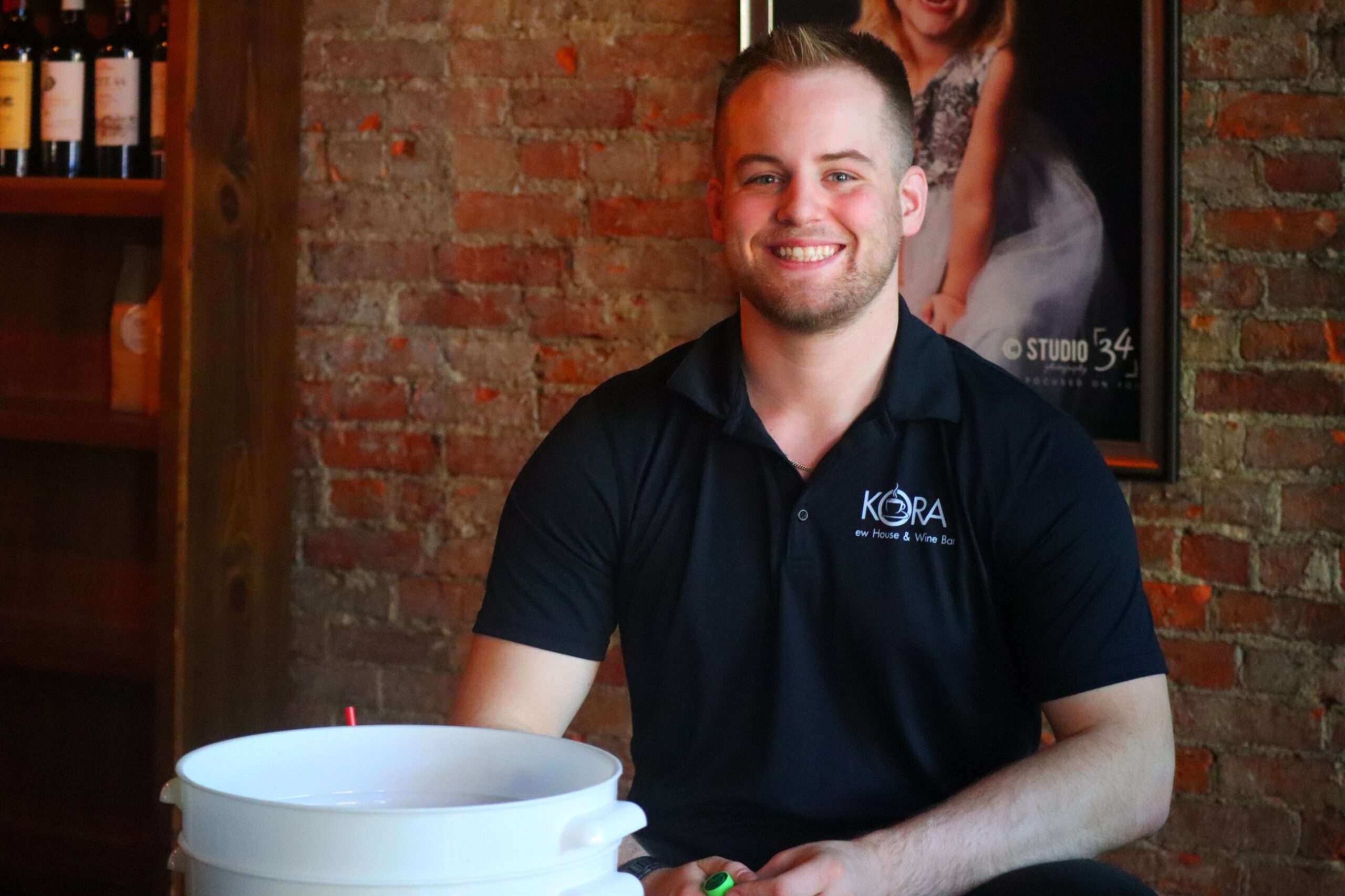 A young man in a black KORA Brew House polo sits behind a white bucket inside Kora Brew House during the Bryan Chocolate Walk.