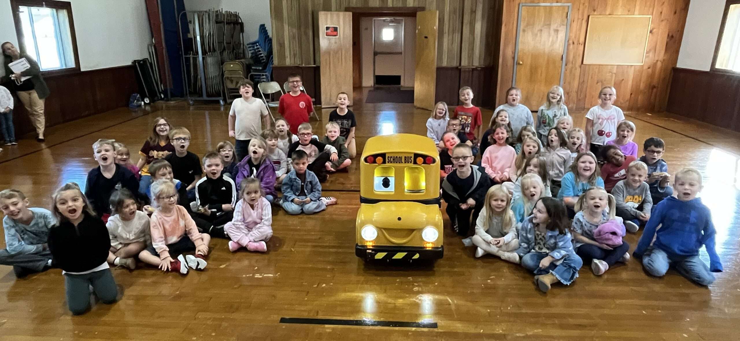 Students sitting on a gym floor around Buster, a small yellow robotic school bus