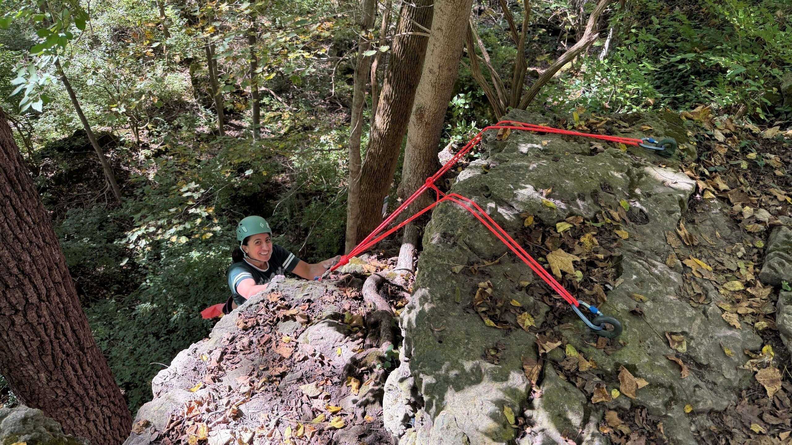 A climber in a green helmet looks up from a rock ledge with red ropes