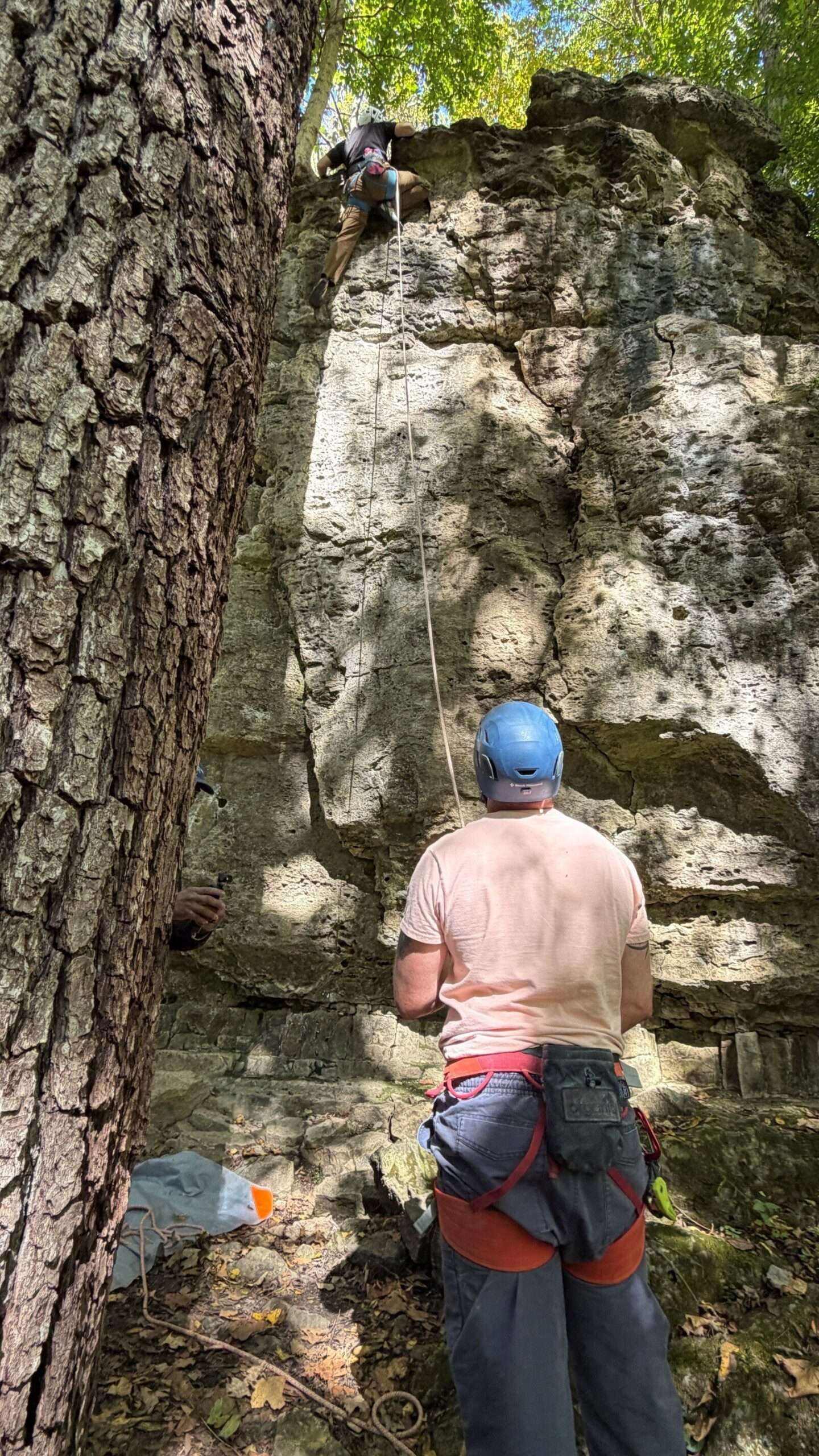 A climber in a blue helmet watches another climber scale a limestone cliff
